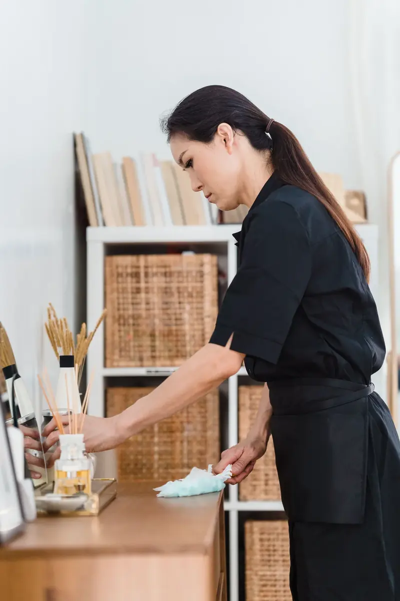 Cleaner dusting a wooden sideboard and styling items during a house clean